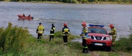 Zdjęcie przedstawia odcinek rzeki Wisły. Przy brzegu stoi 5 strażaków oraz samochód, którym przetransportowano łódź. W oddali łódź ze strażakami. na trzecim planie obszary zielone.