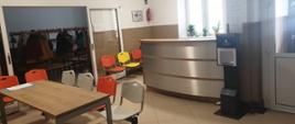 Na zdjęciu znajduje się recepcja PSM, wysoka lada w kolorze srebrnym, widok na szatnię za rozsuwanymi szklanymi drzwiami , stół i krzesła w kolorach szary, pomarańczowy i żółty