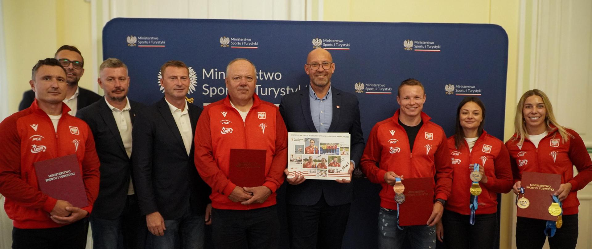 Spotkanie ministra Jakuba Rutnickiego z kajakarzami. Zawodnicy i trenerzy stoją w czerwonych dresach na tle ścianki z logiem ministerstwa. Minister stoi w granatowym garniturze