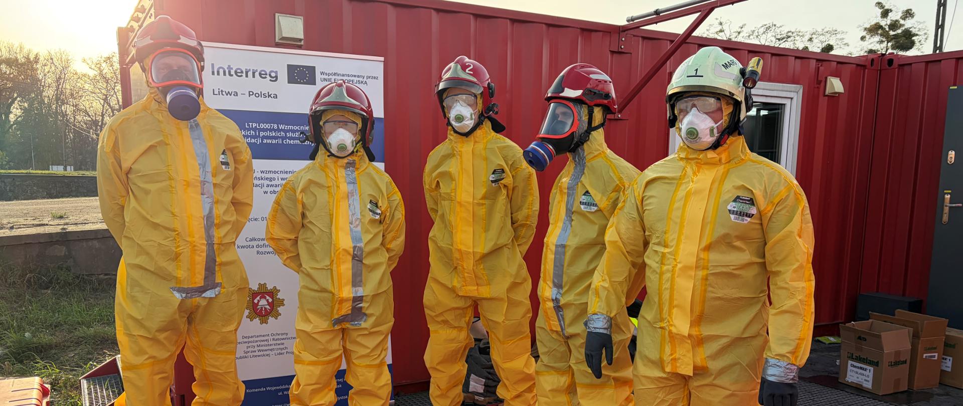 Szkolenia w ramach projektu LTPL00078 "Wzmocnienie współpracy litewskich i polskich służb ratowniczych w zakresie likwidacji awarii chemicznych i ekologicznych" - strażacy w trakcie szkolenia, ubrani w umundurowanie ochronne tychemy, w tle plakaty i banery dot. współpracy 