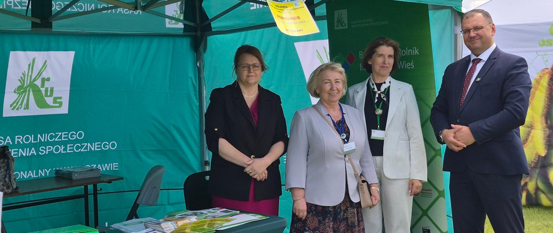 Cztery osoby stojące przy zielonym stoisku informacyjnym KRUS podczas wydarzenia plenerowego. Obok nich znajduje się stolik z materiałami informacyjnymi. 