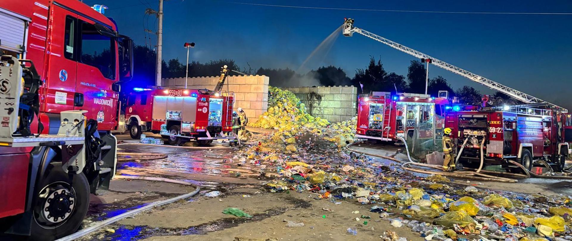 Widok z przodu. Miejsce pożaru - składowisko odpadów. Po lewej stronie dwa samochody gaśnicze oraz prądownik operujący prądem wodnym ze stanowiska na drabinie. Nad sekcją magazynową prądy gaśnicze podawane z samochodu - drabiny, po prawej stronie dwa samochody gaśnicze, a w tle samochód - drabina. Pora wieczorna. 