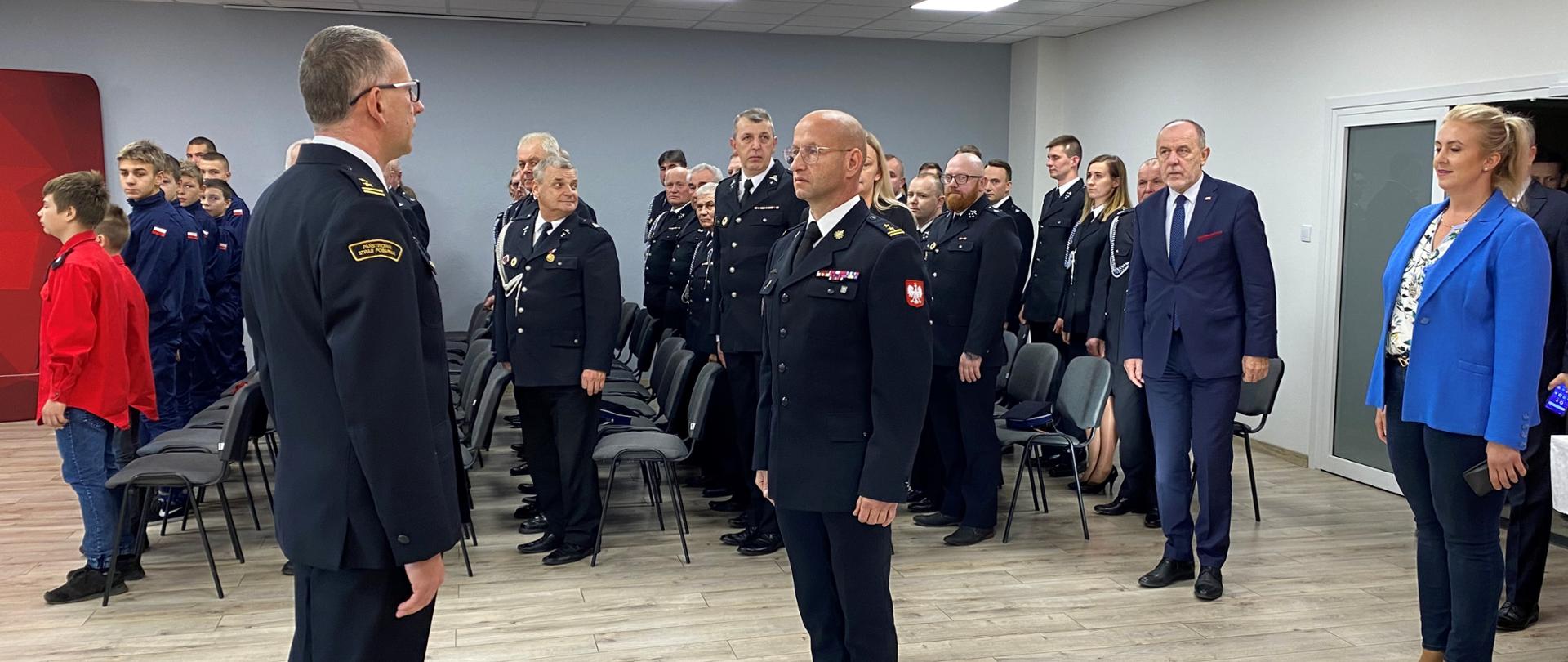 składanie meldunku strażakowi przez drugiego strażaka, w tle na baczność stoją strażacy, oraz kobietaw niebieskiej marynarce i spodniach oraz dwóch mężczyzn w garniturach