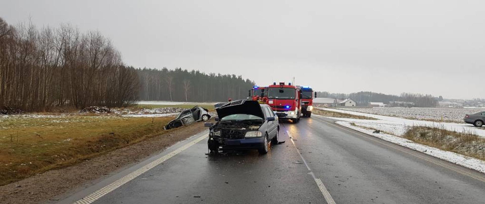 Zdjęcie przedstawia granatowy samochód osobowy z rozbitym przodem stojącym na środku pasa drogowego oraz szary samochód osobowy stojący na kołach w rowie z rozbitym bokiem. W tle widać trzy samochody ratowniczo-gaśnicze. Zdjęcie wykonano z pasa drogowego w pochmurny zimowy dzień.
