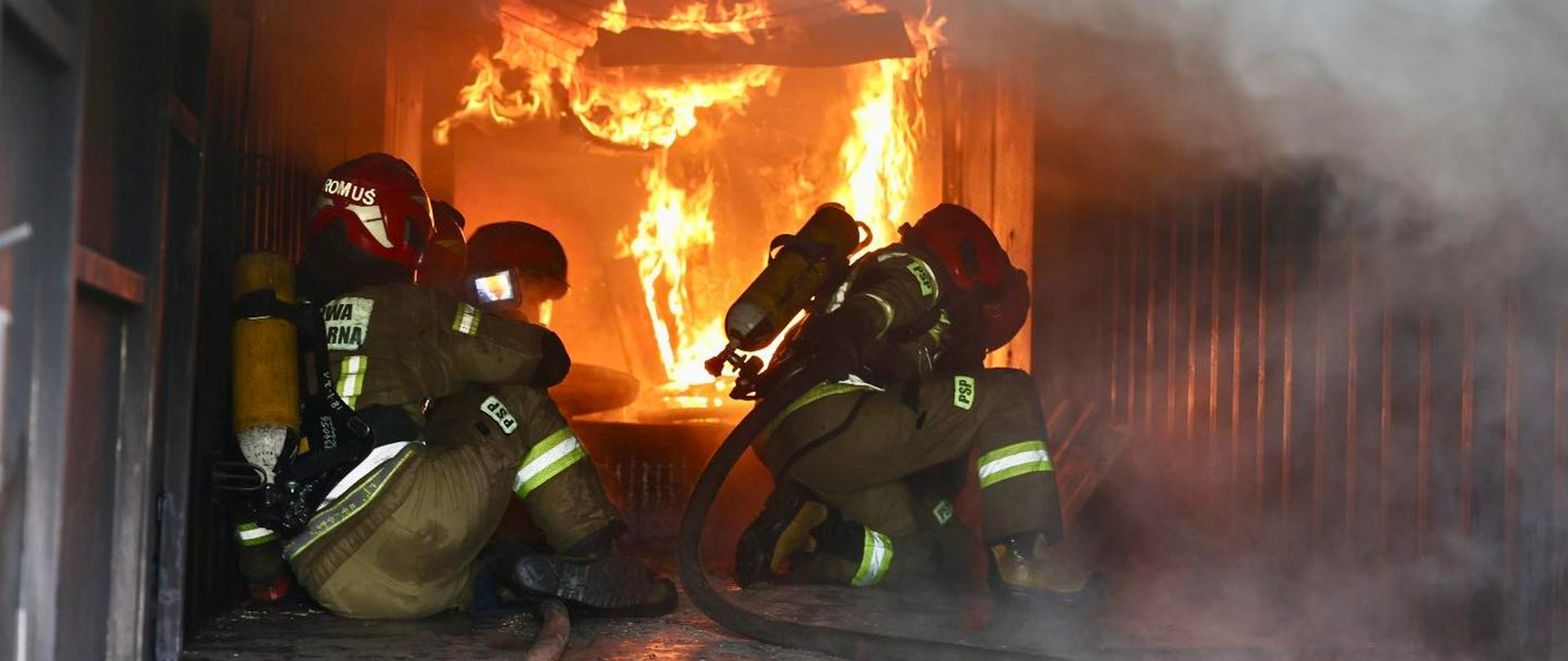 Zdjęcie wewnątrz ciemnego kontenera szkoleniowego, z dużą ilością dymu. Widać trzy osoby ubrane w pełne umundurowanie strażackie, z hełmami i aparatami powietrznymi, kucające na mokrej podłodze. Pośrodku kadru, za strażakami, w głębi pomieszczenia, szaleje intensywny pożar z dużymi płomieniami w kolorach żółtym, pomarańczowym i czerwonym, wypełniający część tylnej ściany. Strażacy są skierowani w stronę ognia, ale ich twarze są zasłonięte przez hełmy i akcesoria. Wzdłuż podłogi, w kierunku ognia, ciągnie się wąż gaśniczy. Dym jest gęsty i wypełnia górną oraz prawą stronę kadru, tworząc zamgloną atmosferę. 