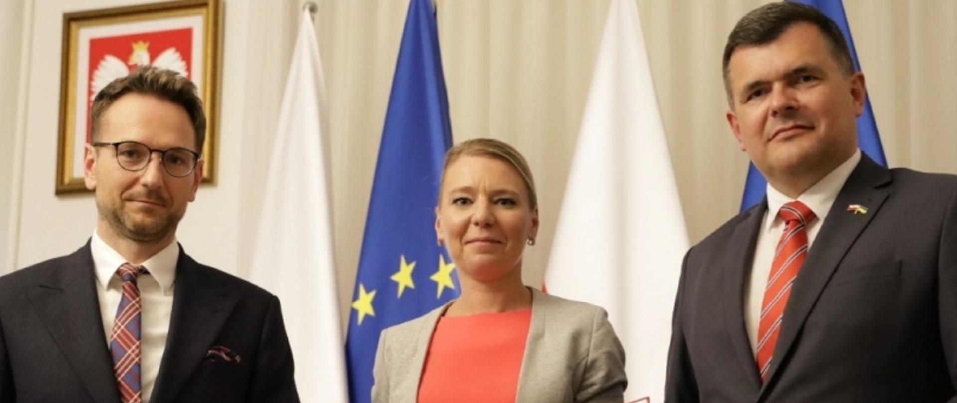 Trzy osoby na tle flag Polski, UE i godła narodowego z białym orłem.