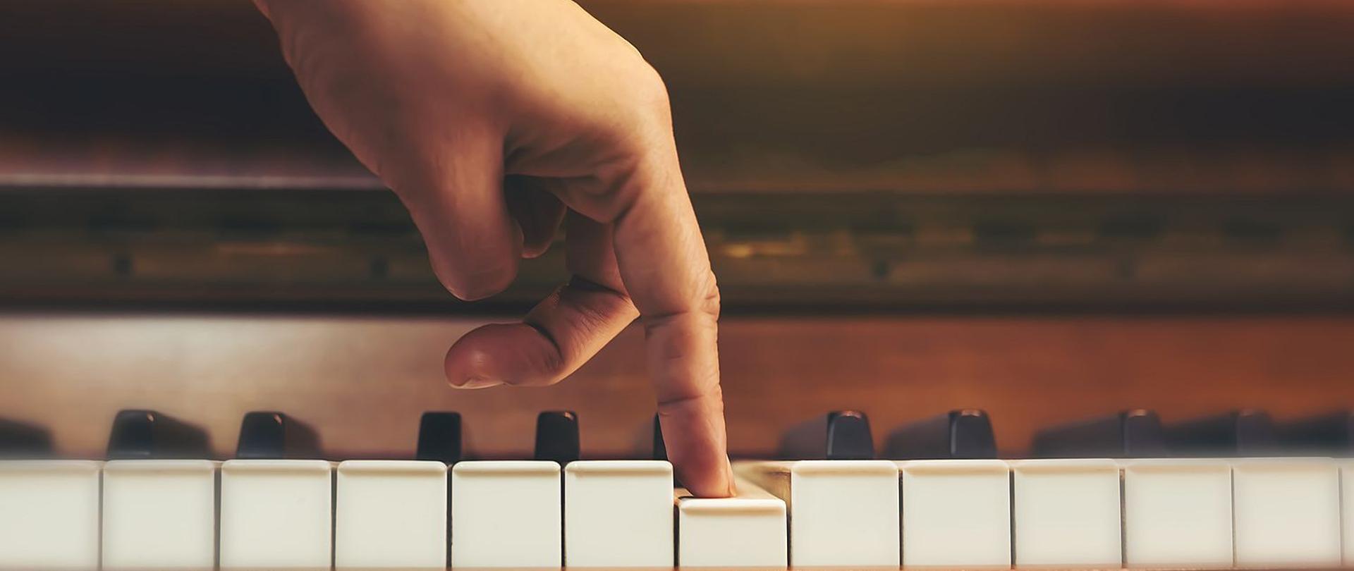Zdjęcie przedstawia rękę wciskającą pionowo jeden klawisz na klawiaturze brązowego pianina.