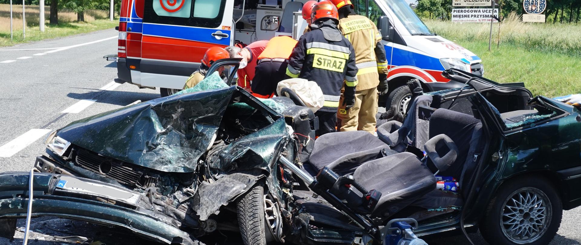 Na miejscu widoczny wrak samochodu osobowego, za nim strażacy oraz ratownicy medyczni wykonujący czynności ratunkowe. W tle widoczna karetka. 