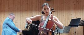 Nastolatka siedząc na krześle na środku estrady gra na wiolonczeli, za nią na pianinie gra mężczyzna.