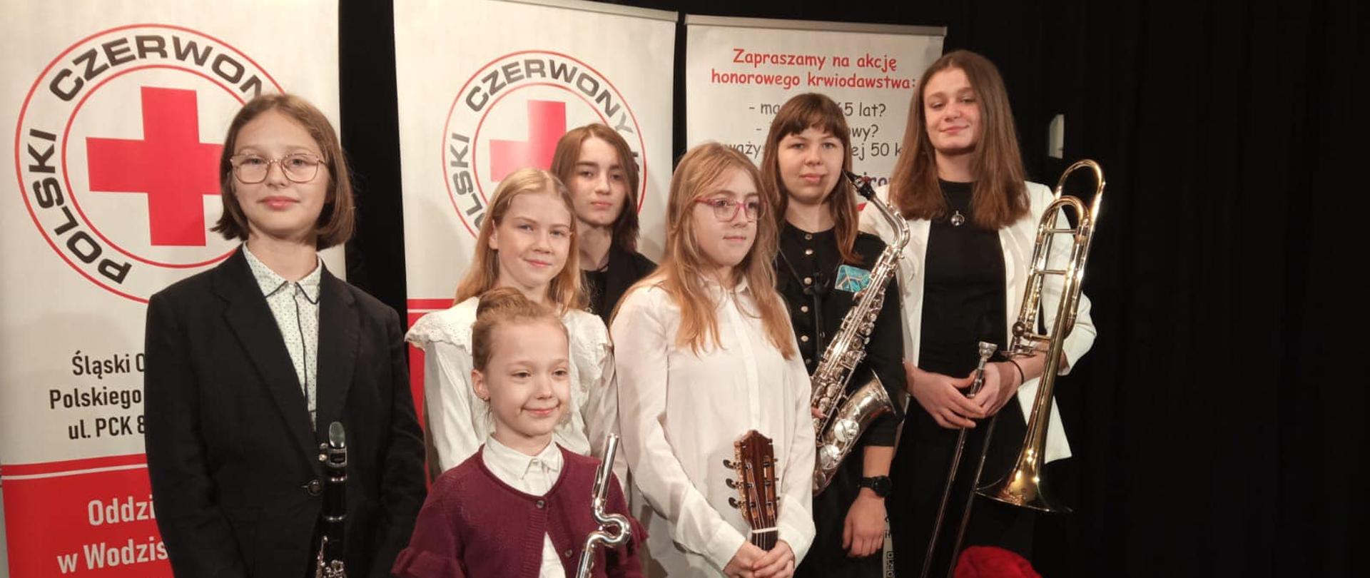 Grupa uczniów stoi na tle plakatów Polski Czerwony Krzyż, maskotka PCK kropelka krwi