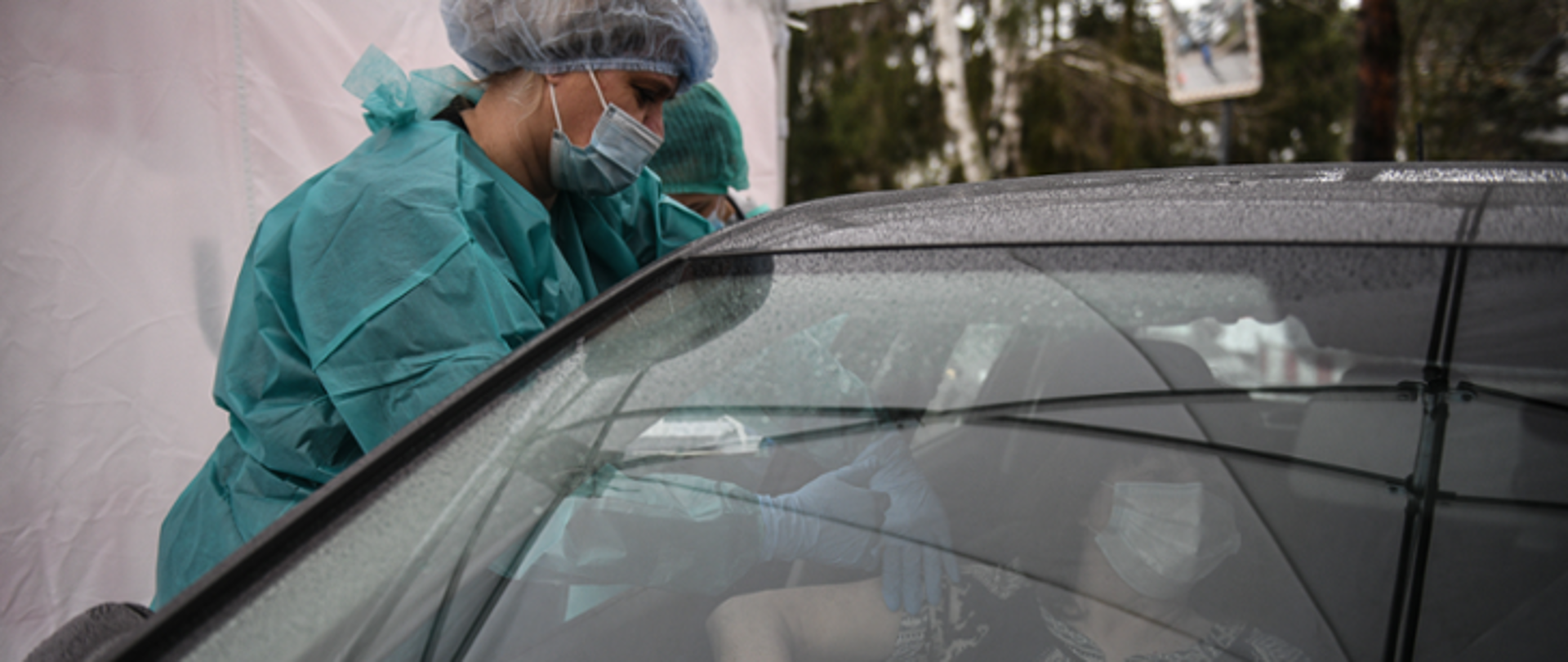 Zdjęcie przedstawia jak wygląda punkt drive-thru, kobieta w kombinezonie szczepi kobietę siedzącą w samochodzie. 