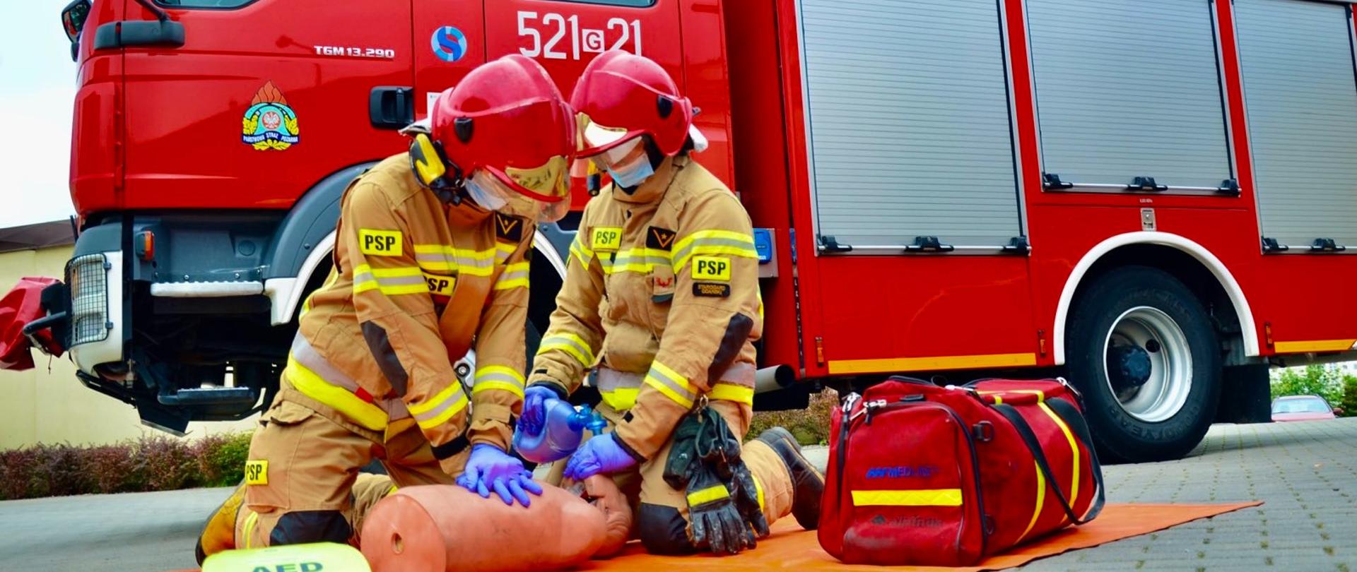 Zdjęcie przedstawia dwóch strażaków podczas ćwiczenia RKO na fantomie. W tle widać samochód pożarniczy