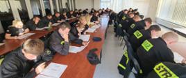Strażacy – ochotniczy w ciemnych ubraniach koszarowych siedzą przy stolikach na sali egzaminacyjnej podczas pisania testu teoretycznego.