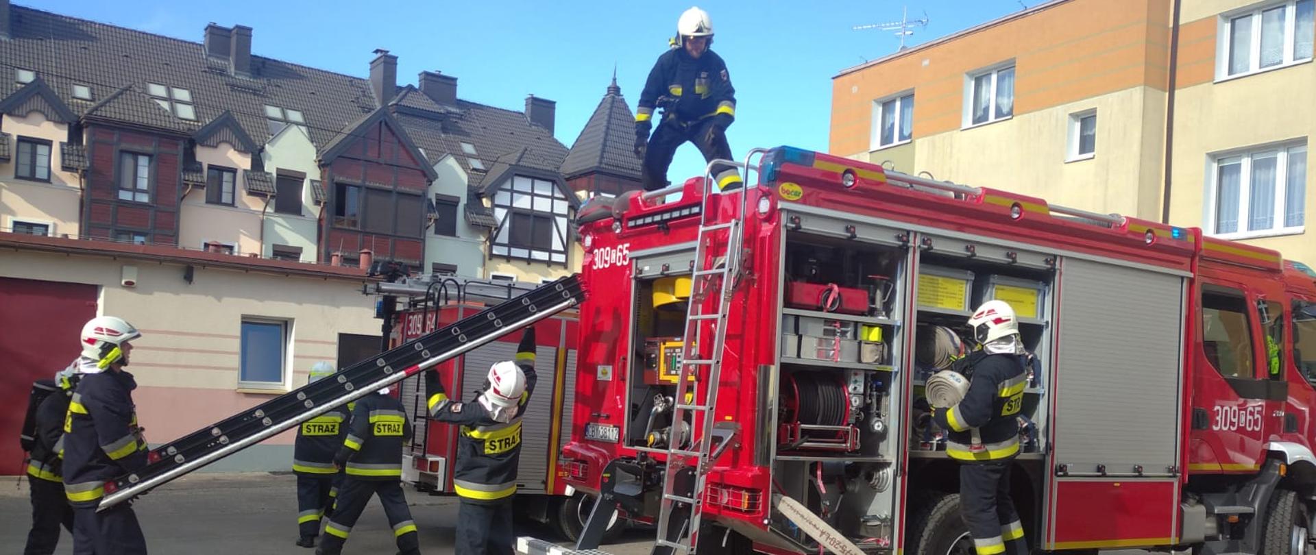 Strażacy w granatowych ubraniach specjalnych biegający przy samochodzie ratowniczo-gaśniczym marki Volvo, widać prawy bok samochodu, trzech strażaków ściąga drabinę z dachu samochodu. 