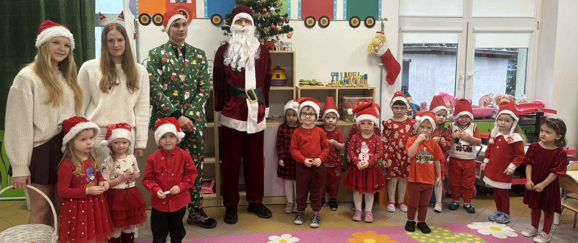 Grupowe zdjęcie dzieci w czapkach Mikołaja, stojących w rzędzie w sali przedszkolnej. Obok nich stoją młodzi wolontariusze w przebraniach - Święty Mikołaj, śnieżynki i elf. W tle widać niewielką choinkę i kolorowe dekoracje z motywem pociągu.