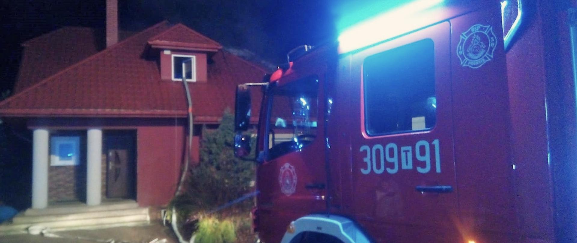 Zdjęcie przedstawia dom jednorodzinny w którym zapalił się dach. Z prawej strony stoi samochód gaśniczy. Wąż pożarniczy leży na chodniku i pociągnięty jest aż do okna w facjacie budynku.