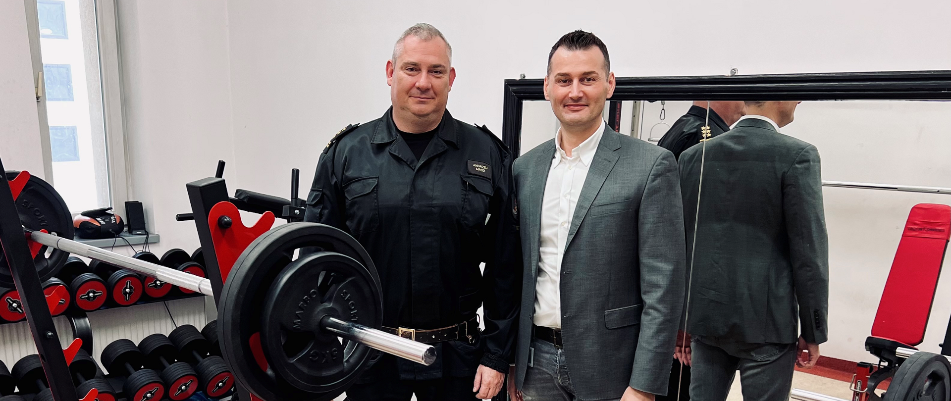 Pomieszczenie siłowni. Na zdjęciu Komendant Powiatowy Dyrektor firmy HUBER+SUHNER. W pomieszczeniu wyposażenie siłowni. Na ścianie duże lustro.