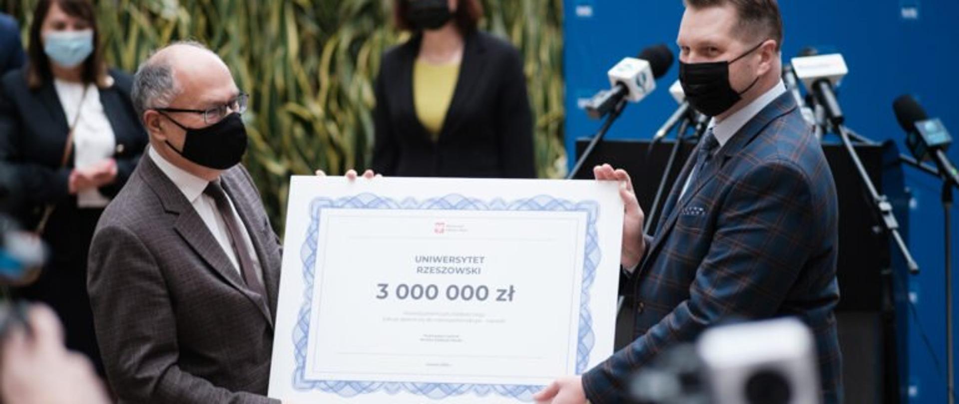 Minister edukacji i nauki Przemysław Czarnek oraz rektor Uniwersytetu Rzeszowskiego Sylwester Czopek trzymają przekazany na rzecz uczelni symboliczny czek z napisem Uniwersytet Rzeszowski 3 000 000 zł. W tle wojewoda podkarpacki Ewa Leniart oraz poseł na Sejm RP Teresa Pamuła