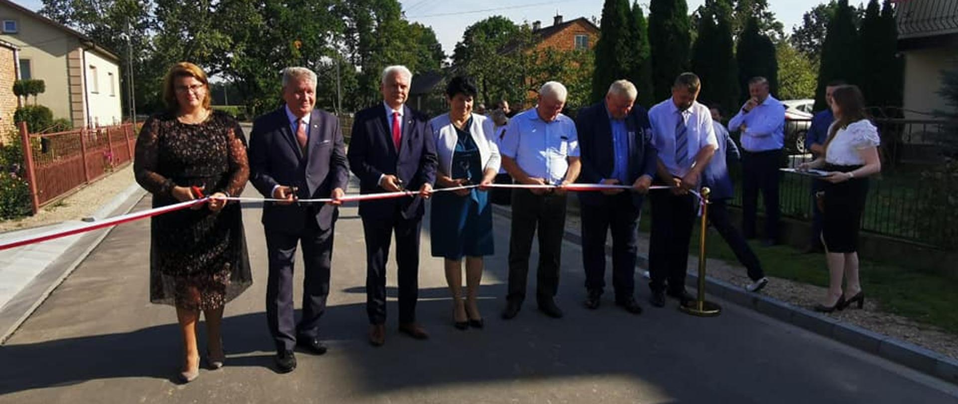 Na zdjęciu znajduje się grupa ludzi w szeregu przecinająca wstęgę, a w tle znajduje się domy