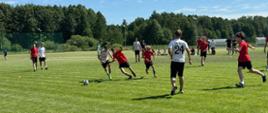 Dynamiczne ujęcie z meczu piłki nożnej rozgrywanego na trawiastym boisku w słoneczny dzień. Kilku młodych zawodników w strojach sportowych (część w czerwonych, część w białych koszulkach) walczy o piłkę w centrum kadru. W tle widać las, pojedyncze lampy oświetleniowe oraz inne osoby obserwujące grę z boku boiska.