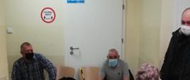 Zdjęcie przedstawia druhów z jednostek Ochotniczych Straży Pożarnych (w umundurowaniu koszarowym i w ubraniu cywilnym), oczekujących w kolejce do punktu szczepień przeciwko COVID-19.