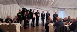 Strażacy w mundurach stoją lub siedzą przy stołach na sali bankietowej
