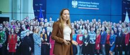 Minister funduszy i polityki regionalnej Katarzyna Pełczyńska-Nałęcz stoi na scenie, za jej plecami ekran z fotografią przedstawiającą pracowników MFiPR