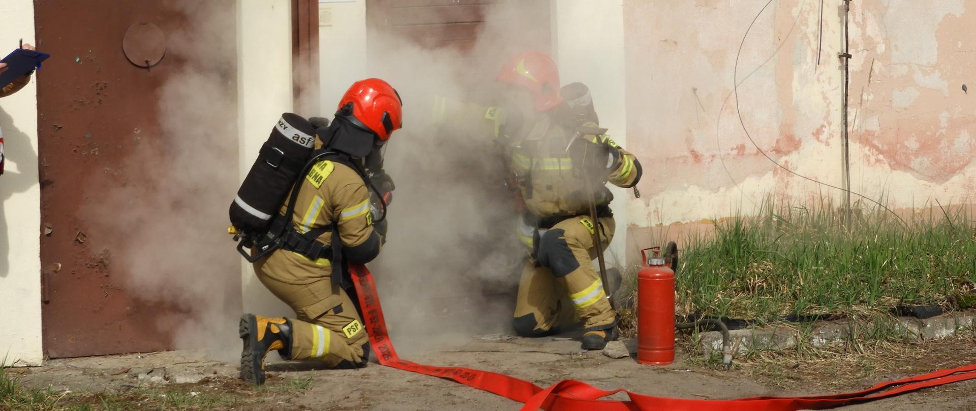 Dwóch strażaków w pełnym umundurowaniu i aparatach ochrony dróg oddechowych pracuje przy zadymionym wejściu do budynku. Jeden z nich klęczy i operuje prądem gaśniczym skierowanym do wnętrza, drugi przygotowuje się do wejścia do środka. Z drzwi wydobywa się gęsty dym. Na ziemi widoczny jest rozwinięty czerwony wąż strażacki oraz gaśnica ustawiona obok wejścia. Budynek ma zniszczoną elewację, a nad drzwiami znajduje się tabliczka z napisem „Krojownia i szwalnia”
