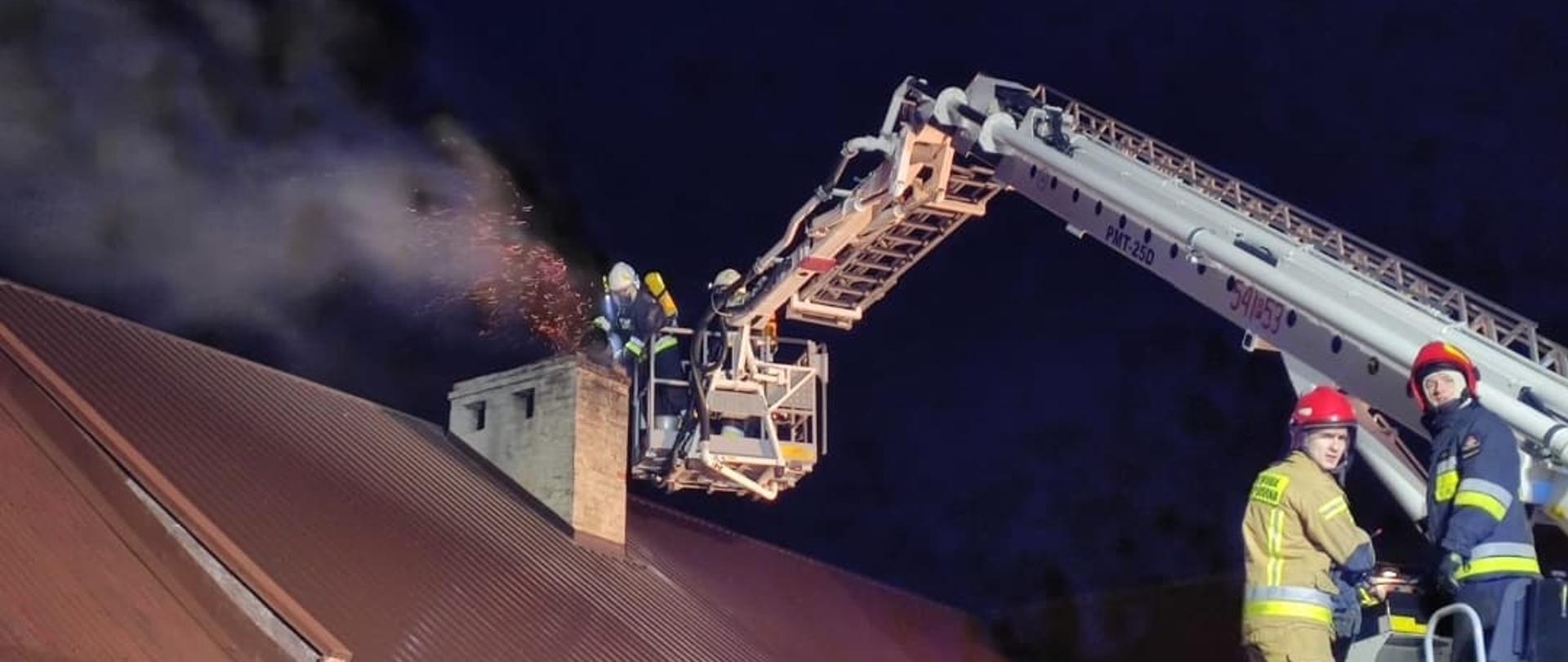 Zdjęcie przedstawia akcję gaśniczą pożaru sadzy. Na pierwszym planie po prawej stronie widać dwóch strażaków ubranych w ubrania specjalne, którzy znajdują się na stanowisku operatora podnośnika koszowego. W dalszym planie widać strażaków w ubraniach specjalnych i aparatach ochrony dróg oddechowych, którzy gaszą palącą się sadze. Znajdują się w koszu podnośnika, który rozstawiony jest nad budynkiem. Poniżej poszycie dachu koloru brązowego. Z komina leci gęsty dym i iskry.
