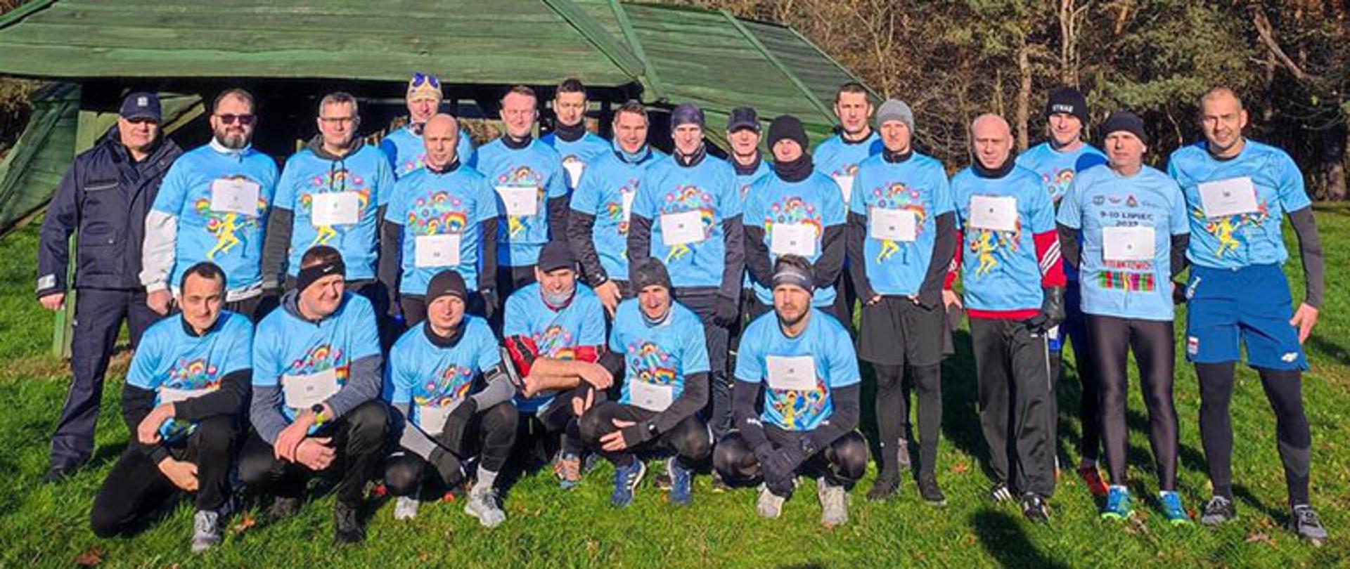 grupa 23 mężczyzn pozuje do zdjęcia pamiątkowego na tle lasu ubrani są w jednakowe niebieskie koszulki z motywem łowickim jeden mężczyzna stoi w ubraniu policyjnym 