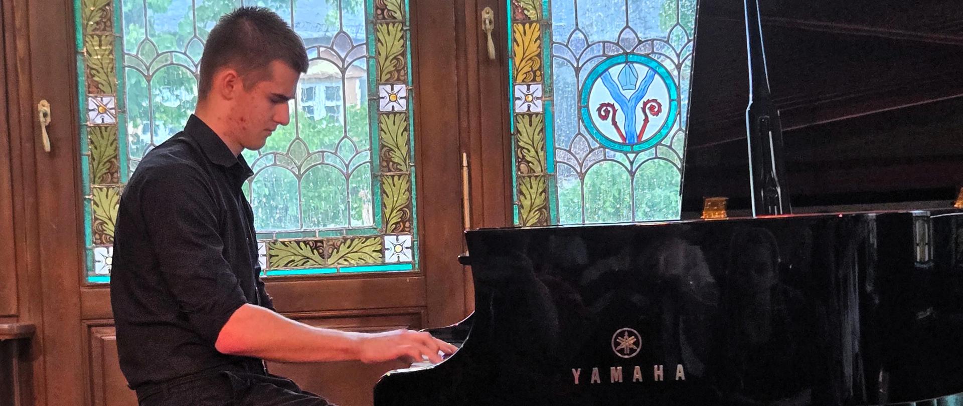 Przy otwartym koncertowym fortepianie siedzi i gra chłopak w wieku nastoletnim ubrany na czarno. W tle można dostrzec okna z kolorowymi witrażami i zielone zasłony.