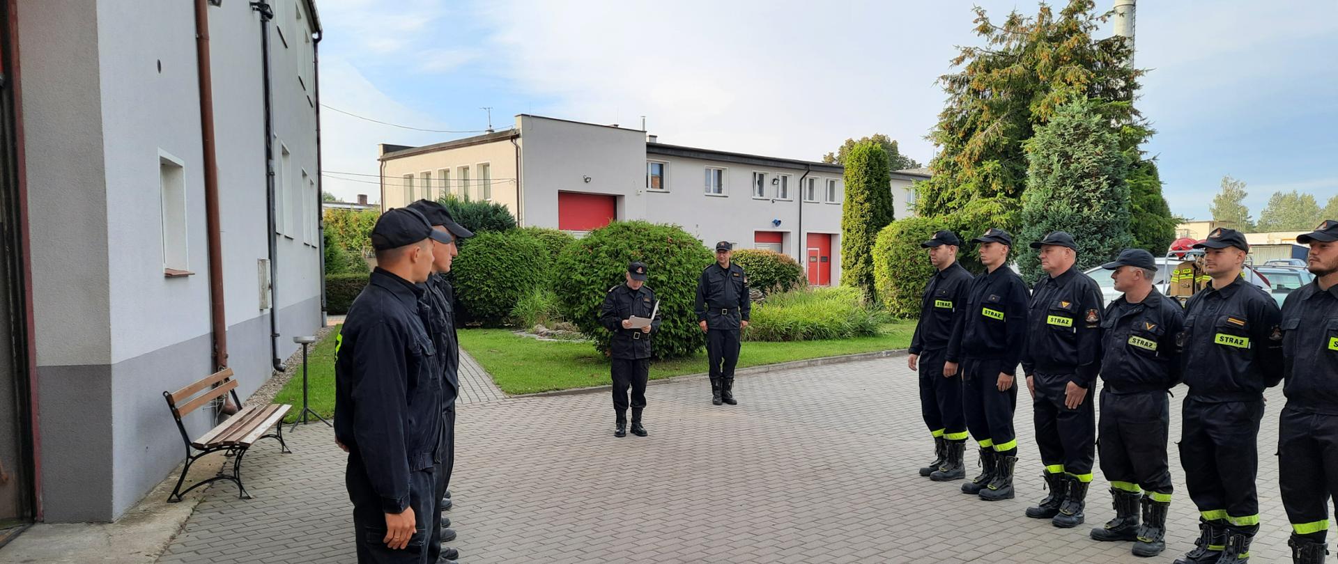 Zdjęcie wykonano w ciągu dnia na placu Komendy Powiatowej Państwowej Straży Pożarnej w Sępólnie Krajeńskim. Na zdjęciu strażacy w mundurach koszarowych stoją na baczność w dwóch szeregach naprzeciw siebie. Na środku stoi Komendant oraz zastępca dowódcy jednostki ratowniczo- gaśniczej ubrani w mundury służbowe. Komendant Powiatowy odczytuje rozkaz. W tle budynek Komendy Powiatowej Państwowej Straży Pożarnej.