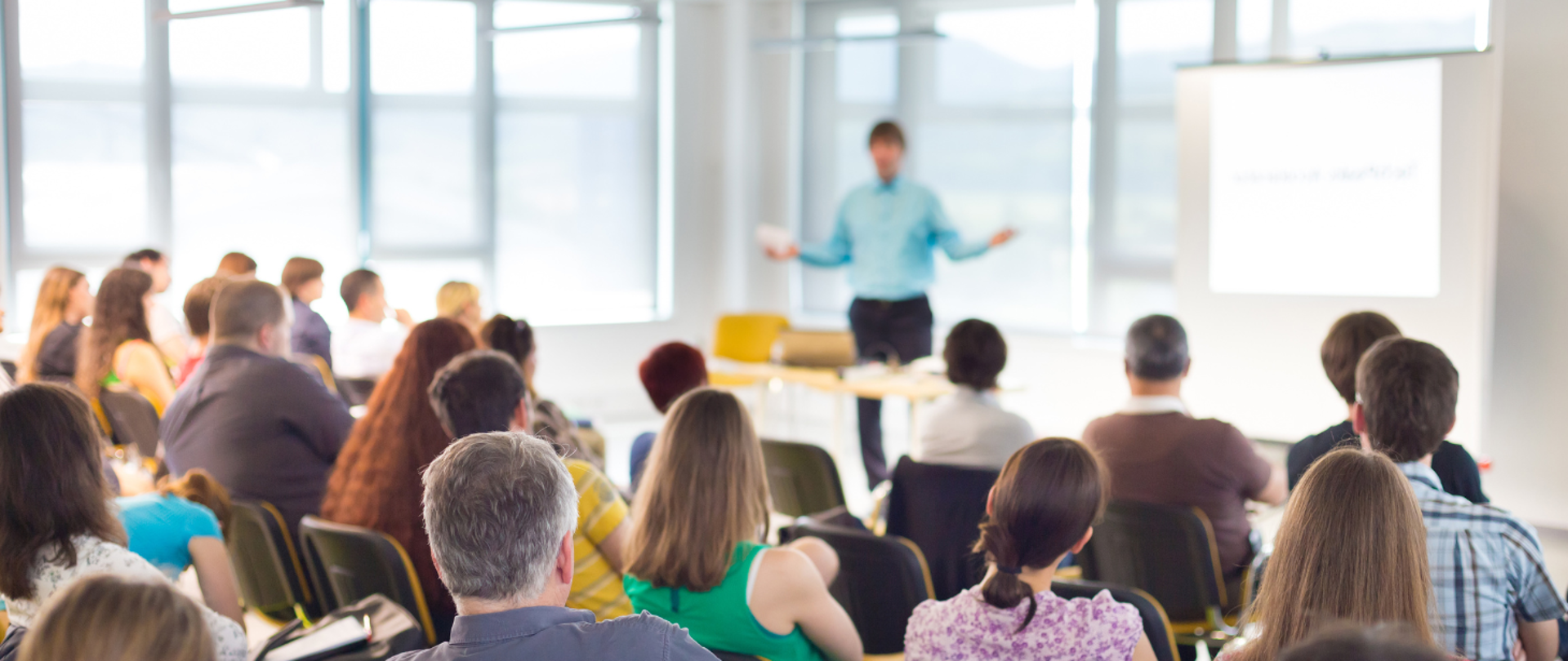 Baner przedstawia jasne, nowoczesne wnętrze sali konferencyjnej z dużymi oknami, przez które wpada naturalne światło. Na pierwszym planie widać grupę osób siedzących na krzesłach w rzędach, zwróconych w stronę prelegenta. W centrum, na tle białego ekranu projekcyjnego, stoi mężczyzna w błękitnej koszuli, prowadzący prezentację. Kolorystyka baneru utrzymana jest w jasnych barwach – dominują odcienie bieli, szarości i błękitu, a także naturalne kolory ubrań uczestników. Baner nie zawiera dodatkowych napisów ani grafik.