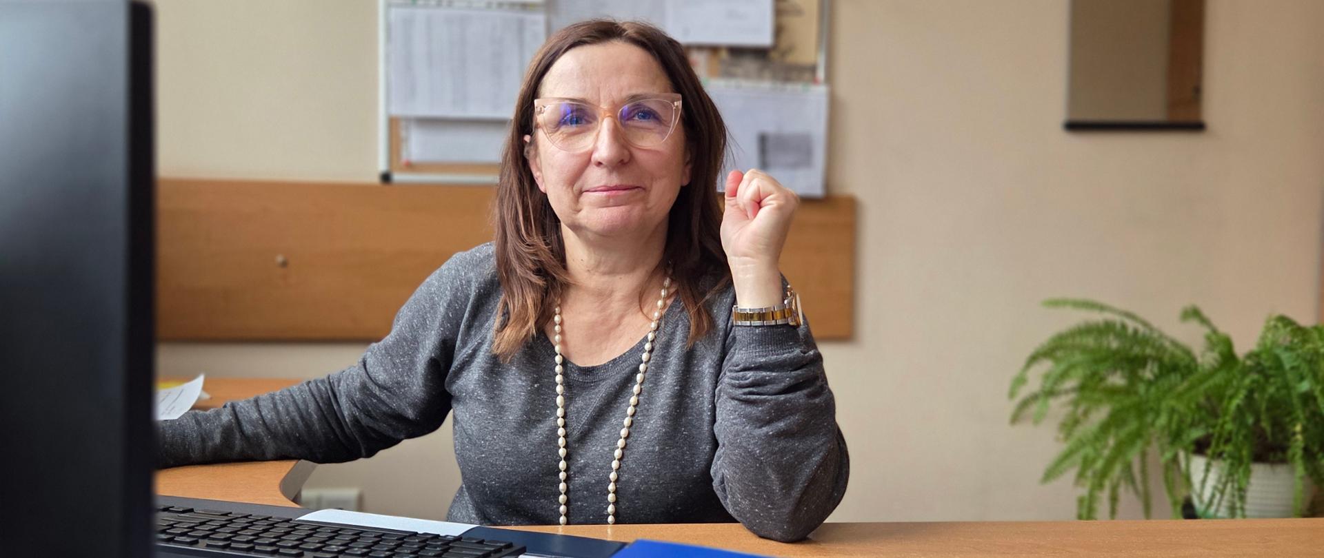Kobieta w okularach siedzi uśmiechnięta za biurkiem, na którym leży niebieski segregator, klawiatura i monitor komputerowy
