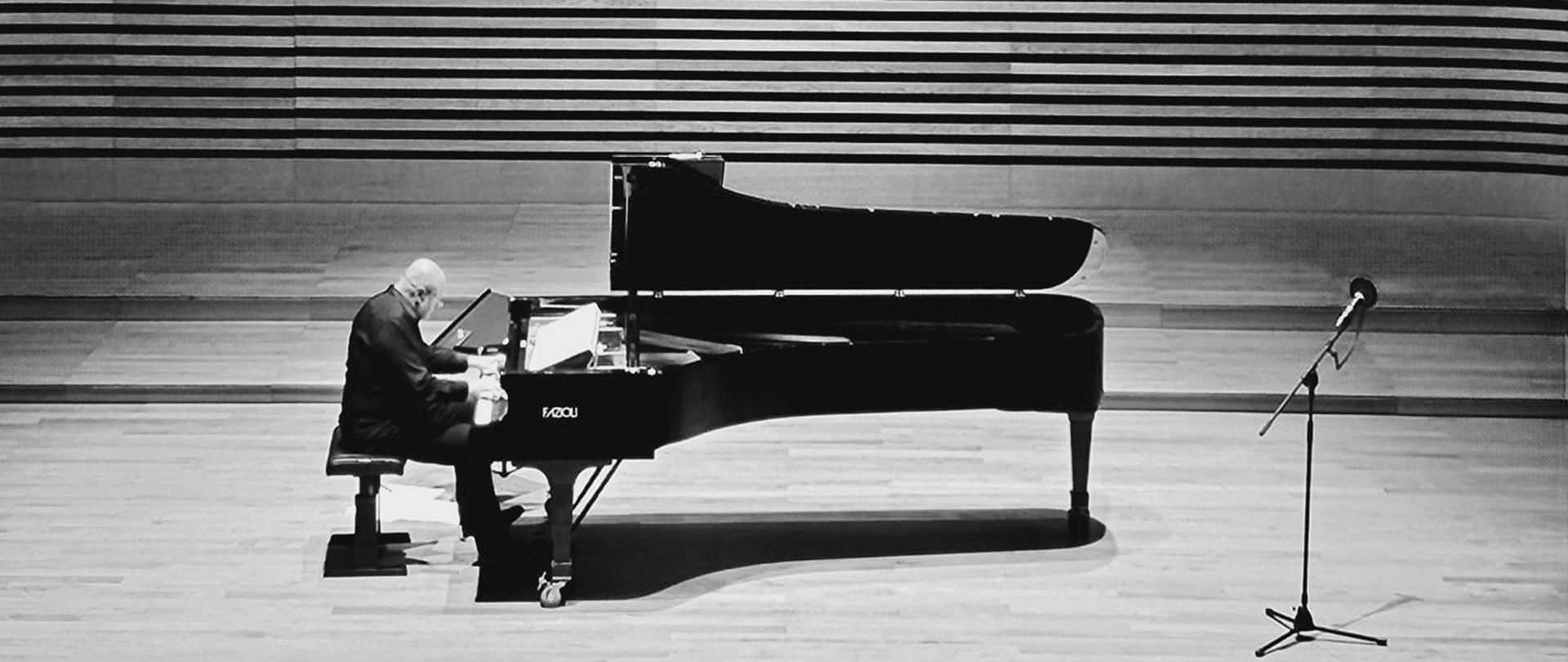 Mężczyzna gra na fortepianie na estradzie sali koncertowej PSM z przodu do tyłu widoczna widownia z publicznością.