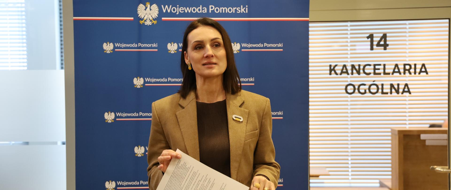 kobieta (wojewoda) stoi w pomieszczeniu, za nią napis kancelaria ogólna