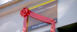 Tablica z nazwą ulicy płk. poż. Zdzisława Filingiera, przy której mieści się siedziba Komendy Wojewódzkiej PSP w Opolu przewinięta czerwoną kokardą przed odsłonięciem. Tablica ma brązowe tło, żółty pas u góry i białe napisy. U dołu na ciemnobrązowym pasie został zamieszczony napis Stare Miasto. Tablica wisi na budynku KW PSP Opole.