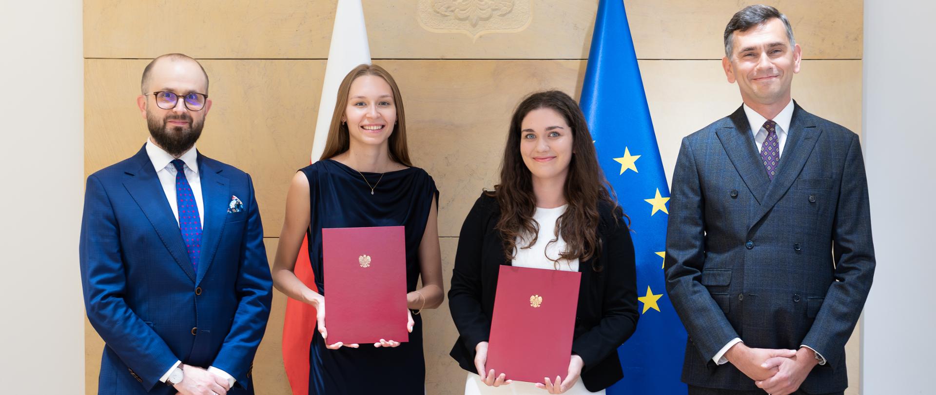 Na zdjęciu są cztery osoby - dwóch mężczyzn i dwie kobiety. Kobiety trzymają bordowe teczki ze złotym orłem. Za nimi są dwie flagi - Polski i Unii Europejskiej. 