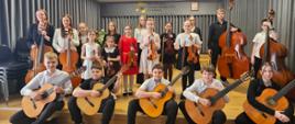 Zdjęcie grupowe przedstawia dziewiętnaścioro młodych uczniów szkoły muzycznej, pozujących na scenie ze swoimi instrumentami. W pierwszym rzędzie, na samym przedzie sceny siedzi pięcioro uczniów z gitarami klasycznymi (czterech chłopców i jedna dziewczyna). Za nimi stoi grupa dzieci trzymających skrzypce, wiolonczele oraz kontrabasy Wszyscy skierowani są w stronę aparatu z uśmiechem na twarzy. W tle widać godło polskie, logo szkoły, szare wytłumiające kotary i czarne krzesła.