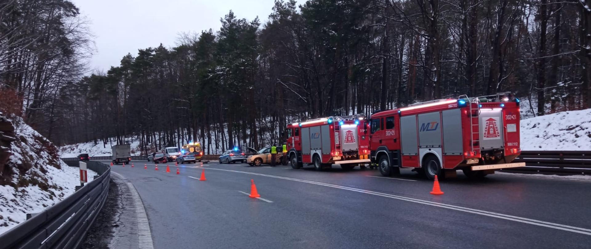 Zdjęcie przedstawia drogę krajową gdzie doszło do wypadku. Wzdłuż drogi stoją kolejno: dwa samochody pożarnicze, uszkodzony pojazd, dwa radiowozy, ambulans i kolejny z uszkodzonych aut. Widać pachołki oznakowania terenu.