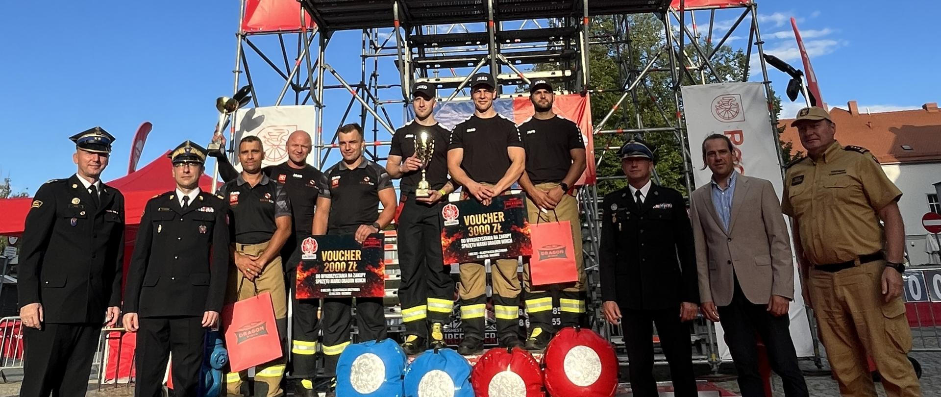Grupowe zdjęcie przed wysoką metalową wieżą treningową udekorowaną banerami sponsorów ("Racibórz OSiR", "Dragon Winch", "GAZ System"). Na podium stoją strażacy w czarnych koszulkach i spodniach bojowych, trzymających puchar i torby z nagrodami oraz vouchery ("2000 zł", "3000 zł"). Po bokach stoją oficjele w mundurach galowych i garniturach. U podstawy wieży leżą niebieskie i czerwone worki treningowe.