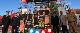 Grupowe zdjęcie przed wysoką metalową wieżą treningową udekorowaną banerami sponsorów ("Racibórz OSiR", "Dragon Winch", "GAZ System"). Na podium stoją strażacy w czarnych koszulkach i spodniach bojowych, trzymających puchar i torby z nagrodami oraz vouchery ("2000 zł", "3000 zł"). Po bokach stoją oficjele w mundurach galowych i garniturach. U podstawy wieży leżą niebieskie i czerwone worki treningowe.