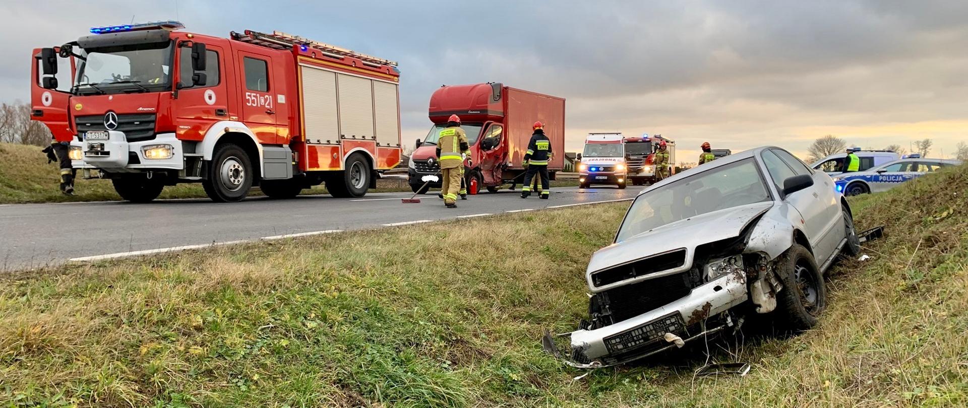 Na zdjęciu po prawej stronie w rowie znajduje się rozbity samochód marki Audi, za nim we wjeździe stoją dwa radiowozy. Po lewej stronie na drodze stoją kolejno samochód GBA, bus biorący udział w kolizji, karetka oraz samochód GCBA. Na zdjęciu znajdują się również strażacy oraz policjanci wykonujący swoje obowiązki