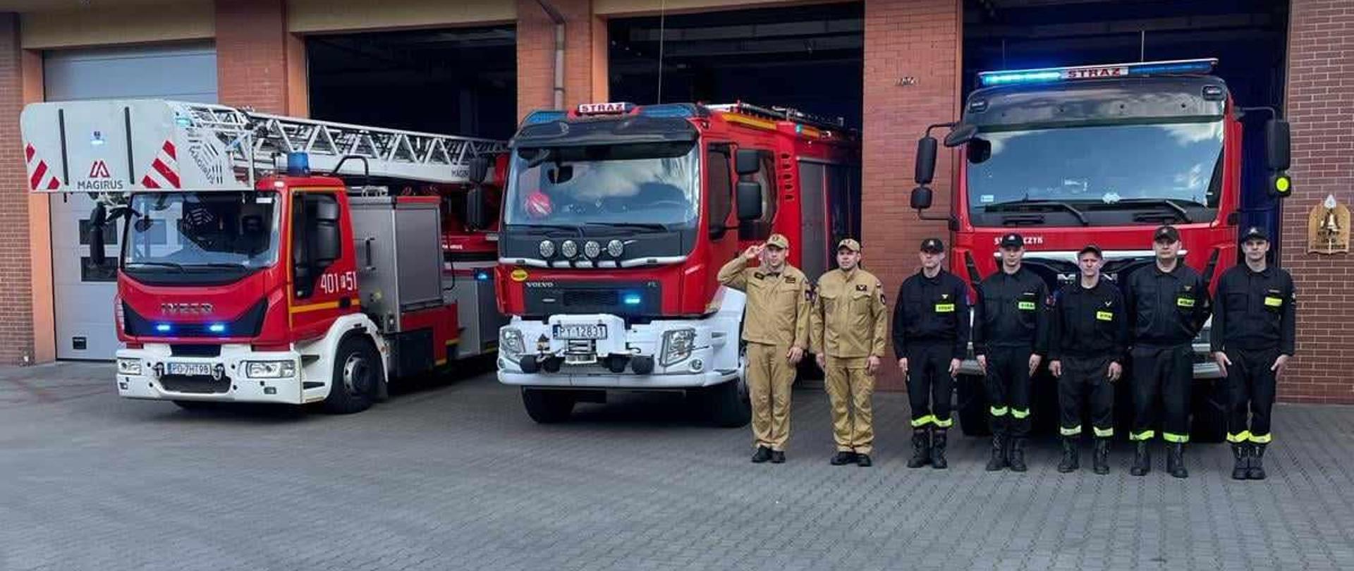 Zdjęcie przedstawia stojących na placu strażaków oddających hołd dla zmarłego strażaka.
W tle wozy strażackie i budynek Komendy.
