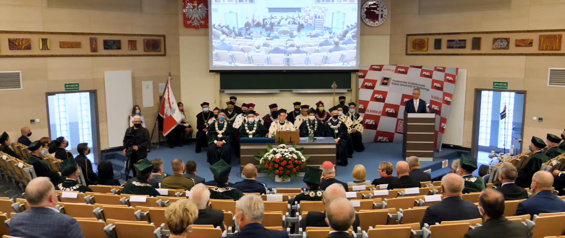 Rzut na aulę akademicką z lotu ptaka. Na pierwszym planie widać rzędy krzeseł i osoby siedzące plecami. Pośrodku auli, na podwyższeniu senat uczelni. Profesorowie ubrani są w togi, łańcuchy i gronostaje. Rektor siedzi przy stole, przed którym znajduje się bukiet biało-czerwonych kwiatów. Po prawej stronie przy mównicy dyrektor Sławomir Adamiec. Na plecami senatu duży telebim. 
