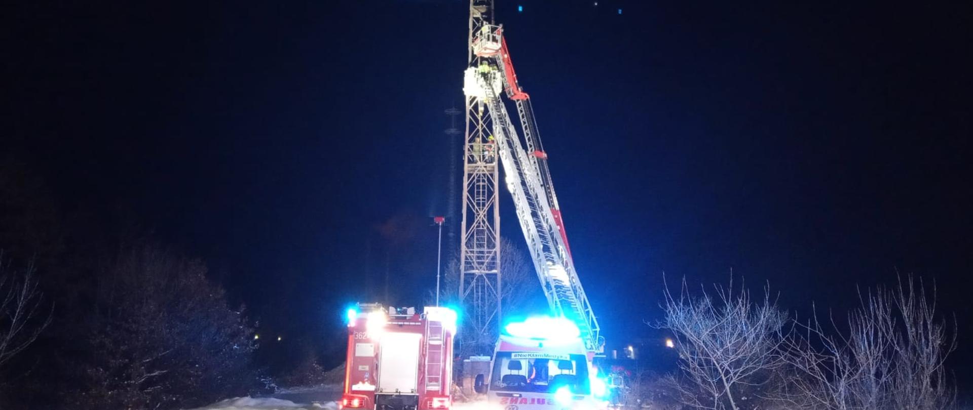 Obraz przedstawia maszt oświetleniowy o konstrukcji z kratownicy. Pora nocna. Na maszcie poszkodowany mężczyzna i ratownicy. Do masztu dostawione dwa kosze z pojazdów pożarniczych. Pod masztem na pierwszym planie widoczny pojazd pożarniczy oraz karetka w włączonymi światłami alarmowymi. Z lewej i z prawej strony widoczne krzaki. Droga dojazdowa pokryta śniegiem.