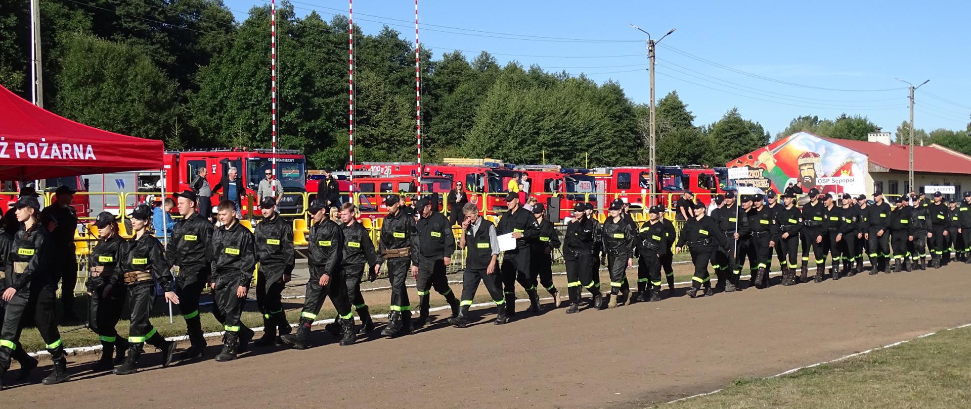 Przemarsz pododdziałów na płytę boiska, w tle pojazdy strażackie