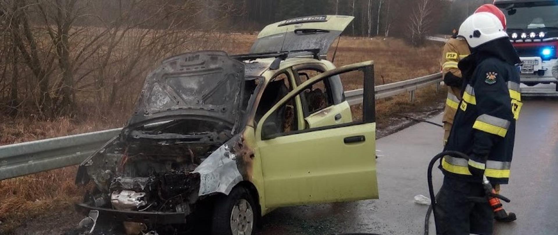 Samochód osobowy stojący na poboczu jezdni po pożarze. Spalona komora silnika oraz wnętrze pojazdu. Obok stoi dwóch strażaków biorących udział w działaniach gaśniczych. W tle samochód ratowniczo-gaśniczy.