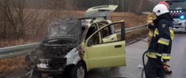 Samochód osobowy stojący na poboczu jezdni po pożarze. Spalona komora silnika oraz wnętrze pojazdu. Obok stoi dwóch strażaków biorących udział w działaniach gaśniczych. W tle samochód ratowniczo-gaśniczy.