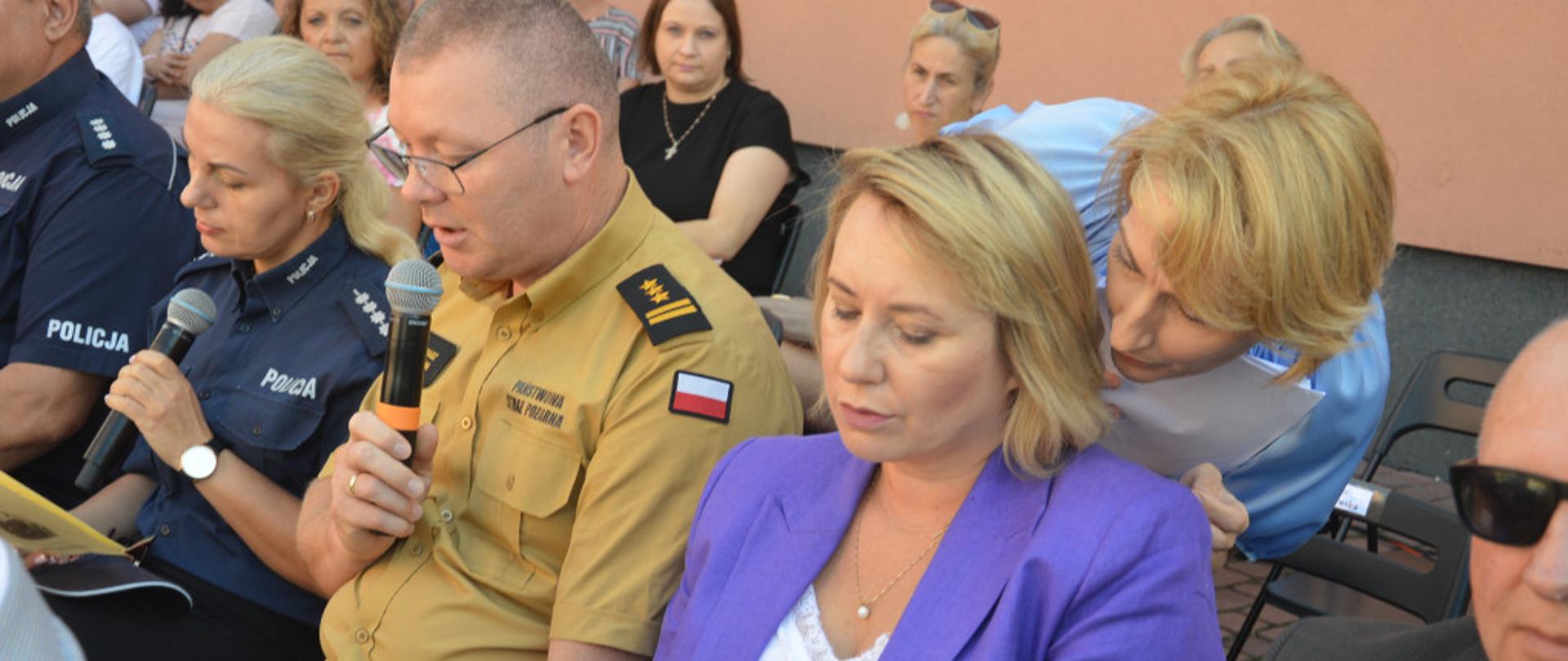 Na zdjęciu widać zaproszonych gości "Narodowego czytania" siedzących na krzesełkach na placu przed budynkiem przed budynkiem Zespołu Szkół Technicznych w Kolnie. Na zdjęciu między innymi jest zastępca komendanta powiatowego PSP w Kolnie st. bryg. Tomasz Sielawa.